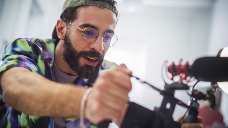 Focused Young Male Cinematographer With Beard And Glasses Adjusts Camera During An Indoor Shoot, Capturing Essence Of Modern Filmmaking And Creative Advertising In A Professional Setting.
