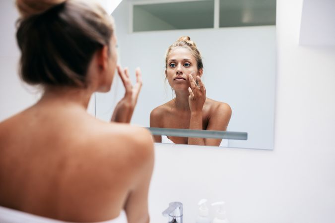 Woman in bathroom looking in to mirror and applying cream on her face