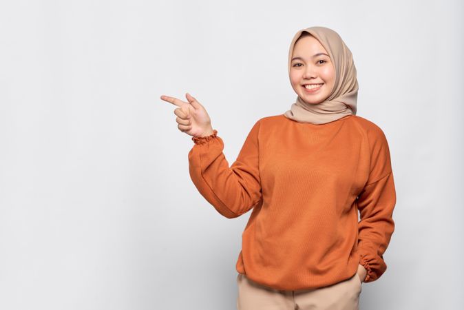 Merry Muslim woman in headscarf and orange sweater pointing to her side