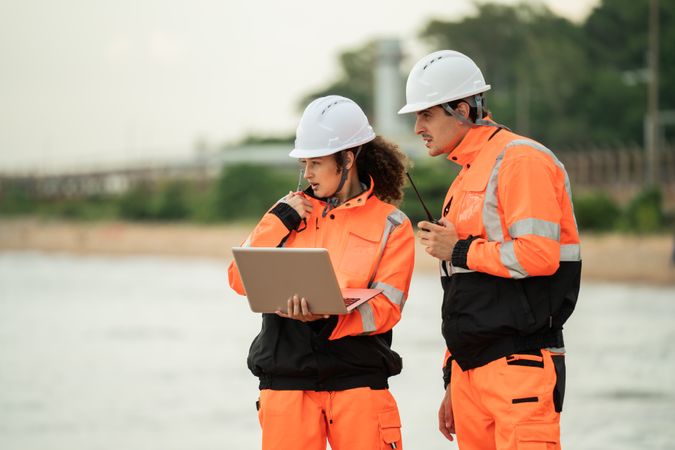 Two engineers wearing safety gear are working at an industrial site near spherical gas storage tanks. Using laptop and walkie talkie to coordinating field operations in a petrochemical environment