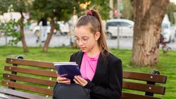 Girl reading book while sitting on park bench.