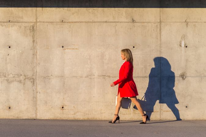 Businesswoman wearing red jacket walking on the street with shadow on cement wall