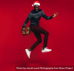 Young Black man wearing Santa hat jumping with gift box over red background 5aAQP5