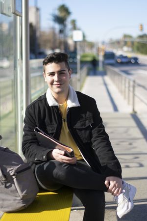 Young student sitting at the bus stop holding a notebook while looking at the camera with a smile