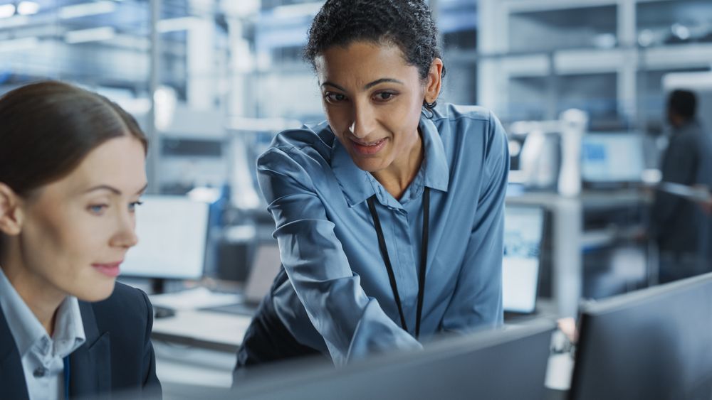 White Female Engineer And Hispanic Product Manager Using Desktop Computer And Talking At Automated Factory With Robotic Hands.
