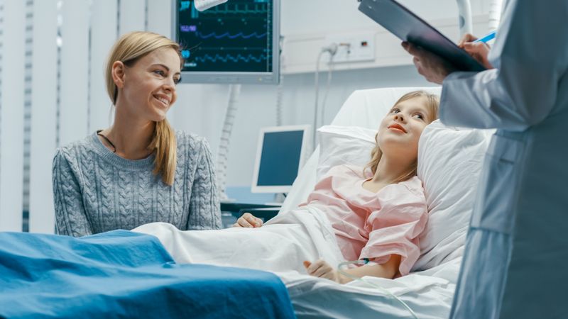 Recovering Little Girl Lies on a Bed In the Hospital, Friendly Doctor With Clipboard Asks Where it Hurts, Mother Sits Beside Bed.