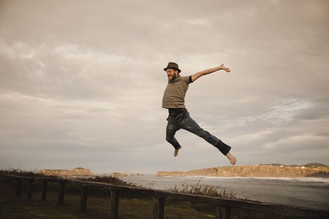 Young man in hats having fun on seat on coast near waving sea and cloudy sky
