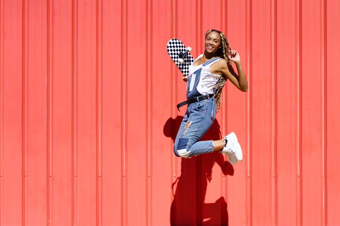 Woman smiling and jumping in front of a red wall with checkered skateboard