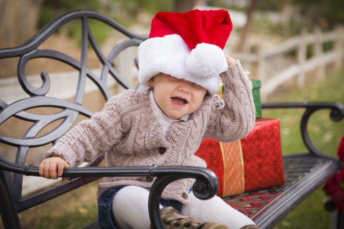 Young Child Wearing Santa Hat Sitting with Christmas Gifts Outside.