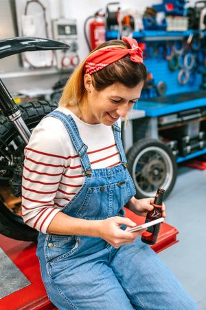 Mechanic woman looking phone and holding beer sitting in front of motorcycle on factory
