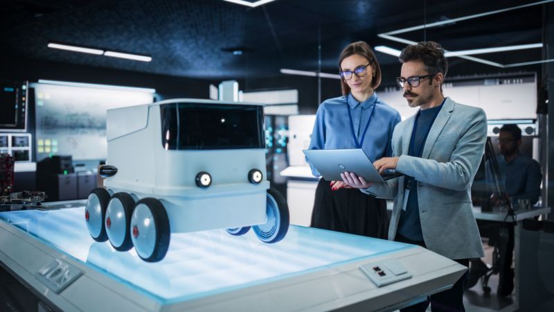 Portrait of Two Colleagues Working in a Research Facility, Using Laptop Computer to Develop a Futuristic Robot for Autonomous Deliveries. Manufacturing Specialist Doing a Demonstration to Team Manager