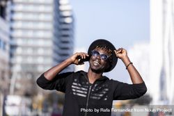 Happy Black man wearing hat & sunglasses standing on the street talking on cellphone 4Bavze