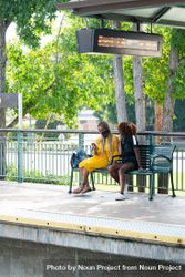 Two women sit outdoors on bench on train platform looking at schedule on mobile phone 5aXdG0
