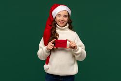 Girl in Santa hat presenting red holiday envelope.