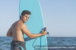 Male surfer holding blue board standing in front of the sea and looking back at camerawater bY3Y65