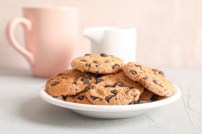 Plate with tasty chocolate chip cookies and blurred cup of milk on gray background, closeup