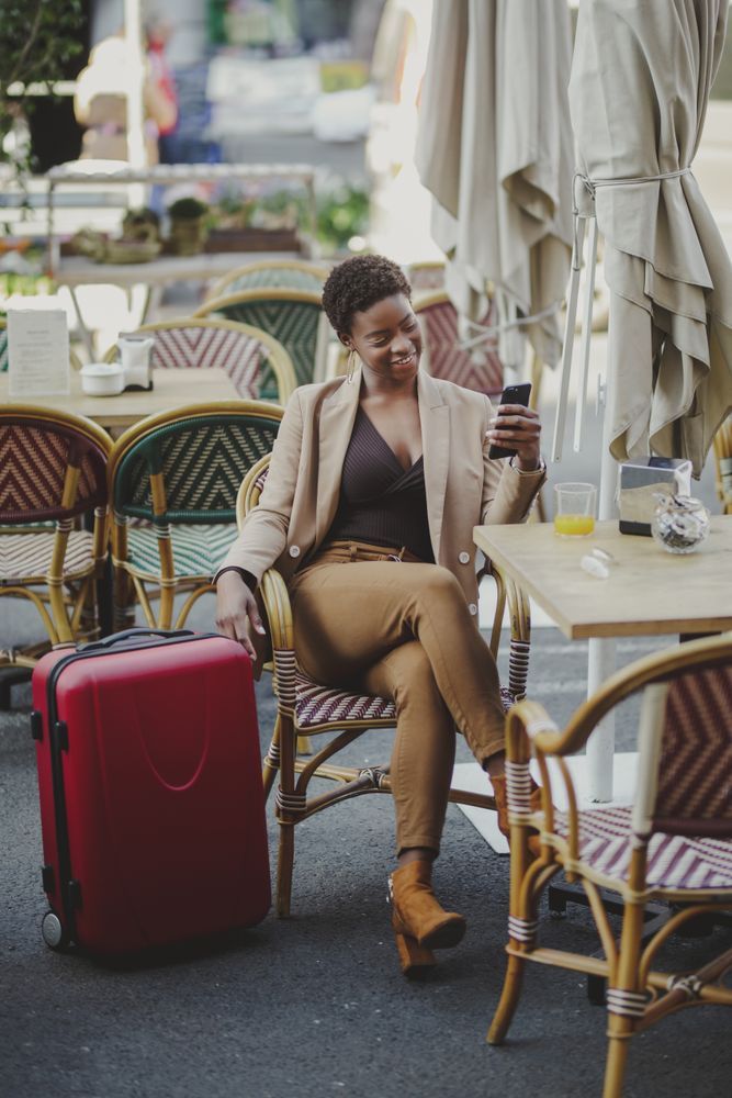 Smiling Black woman with smartphone sitting at table near luggage in cafe