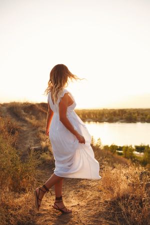 Blonde female in ethereal summer dress turning around to look at lake