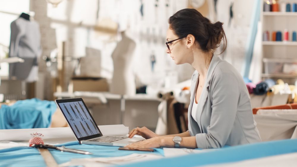 Woman fashion, designer,  Sitting at Her Desk Looking at Sketches on a Laptop, Taking Notes.