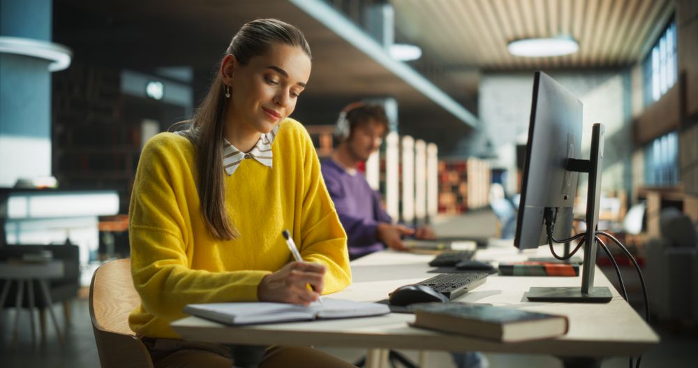 Student Writing a Scientific Essay on a Desktop Computer and Taking Notes in a Paper Notebook. Young Stylish Female Studying in a Public Library with Other People