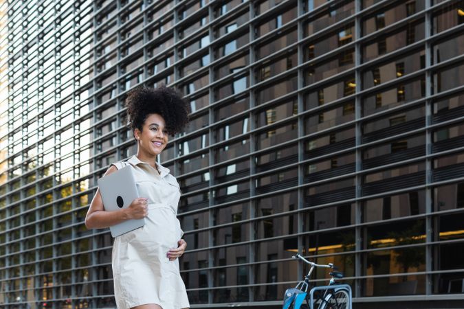 Smiling Black woman with laptop coming out of office building