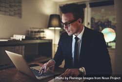 Man in suit and glasses working on his laptop and documents at night 0WrR1b