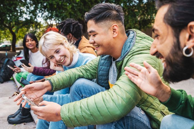 A group of diverse friends laugh and share content on a smartphone while sitting together in an city party