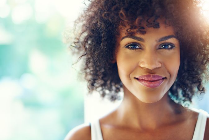 Female with curly hair looking at camera and half smiling on blurry leafy background, copy space