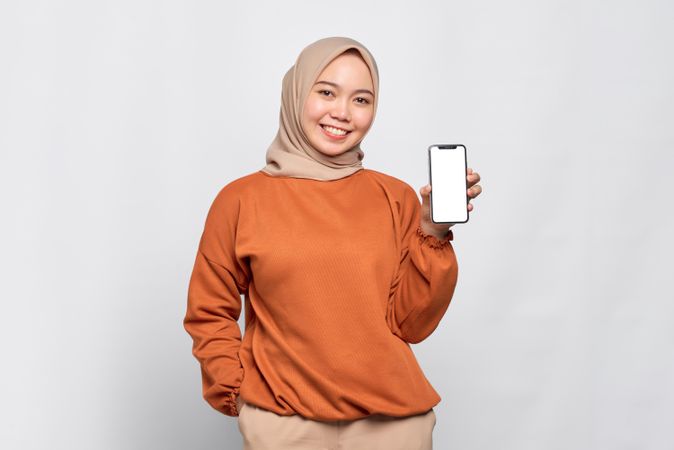 Happy Muslim woman smiling with hand in pocket presenting smart phone with mock up screen