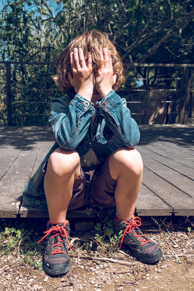 Upset boy sitting on wooden boardwalk and covering face
