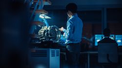 Professional Automotive Engineer in Glasses is Working on Transmission Gears in a High Tech Innovative Laboratory with a Computer Screens.