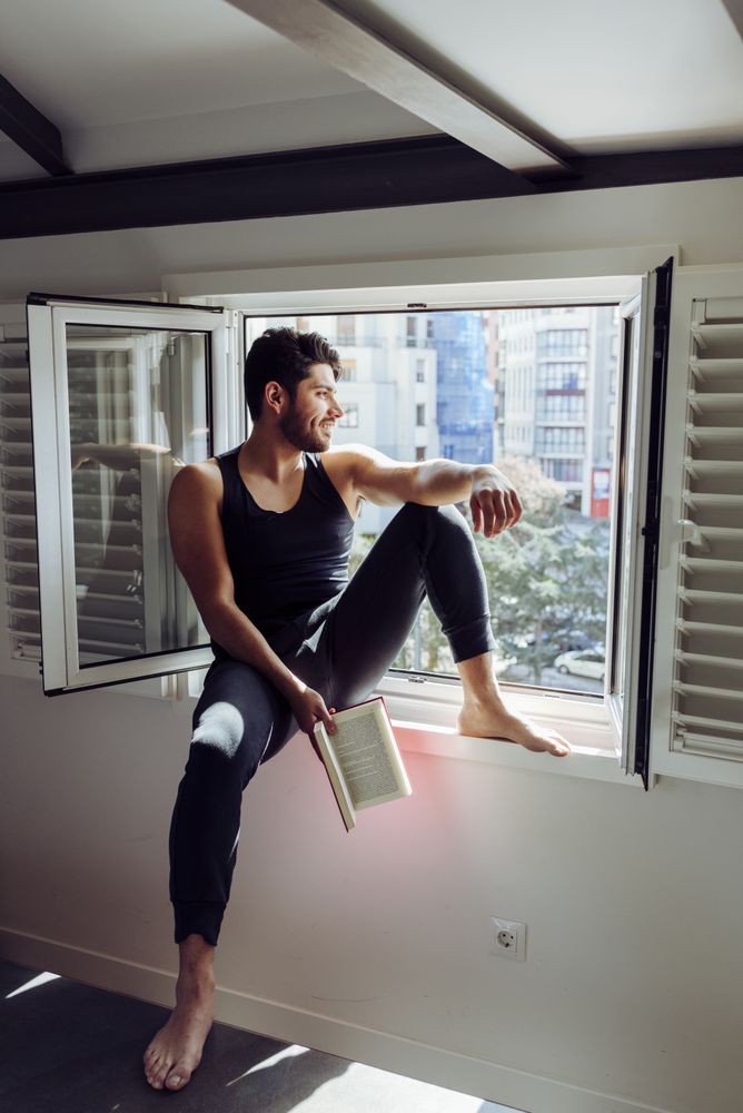 Young male sitting on window and reading book