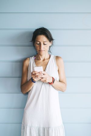 Portrait of woman in summer dress checking phone while leaning on wood wall