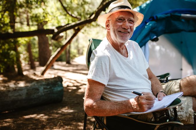 Smiling older man sitting in front of a tent and writing in a book