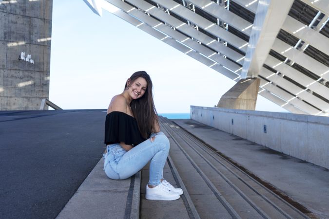 One happy beautiful woman in long hair sitting on staircase at promenade while smiling and looking at camera
