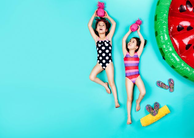Twin girls in swimwear lying down on floor holding colored pineapple with slippers