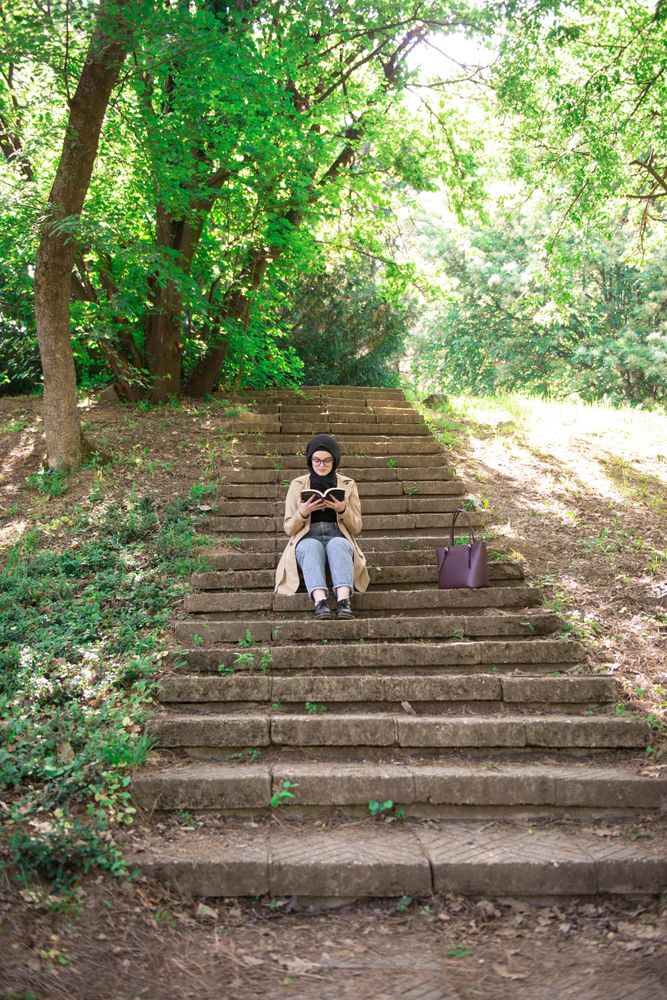 Woman in headscarf reading on park stairs, vertical
