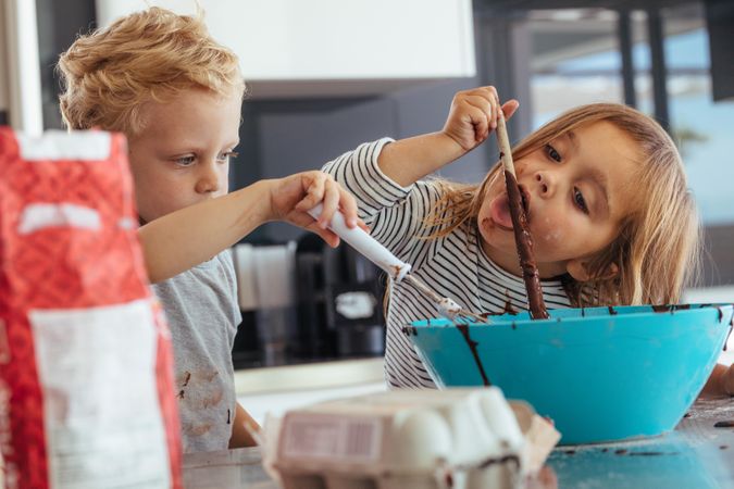 Little kids mixing batter in a bowl for baking, with girl licking a spoon
