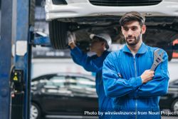 White male mechanic in blue suit holding wrench at auto garage standing under lifted vehicle bY2AG0
