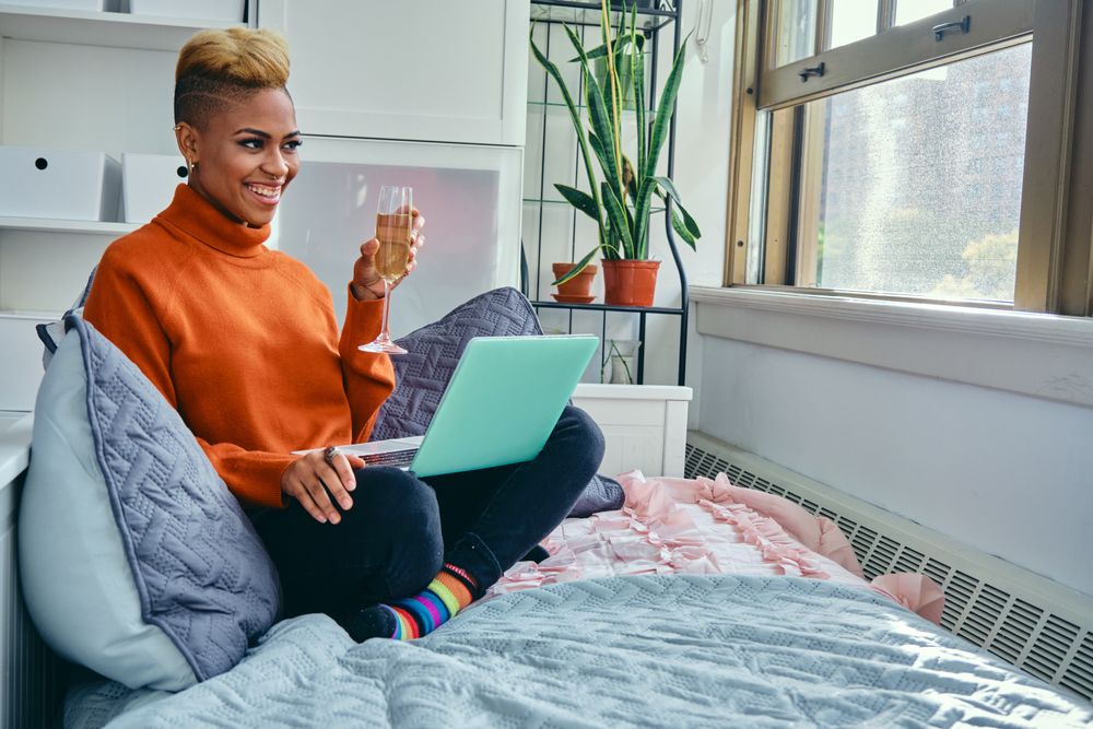 Happy Black woman smiling at a laptop with champagne flute in light filled living room