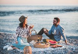 Couple smiling holding wine glasses over summer picnic by the ocean