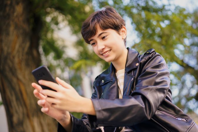A young person joyfully engaged in using their smartphone while seated in urban greenery