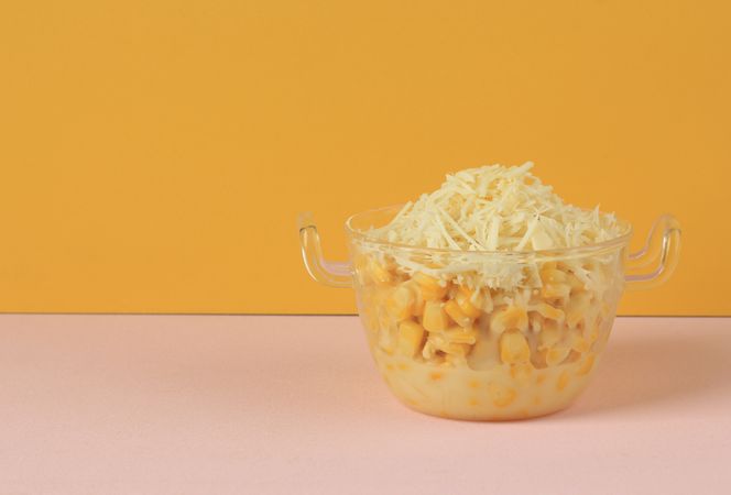 Side view of clear bowl of Indonesian sweet corn dessert with condensed milk and grated cheese on colorful background