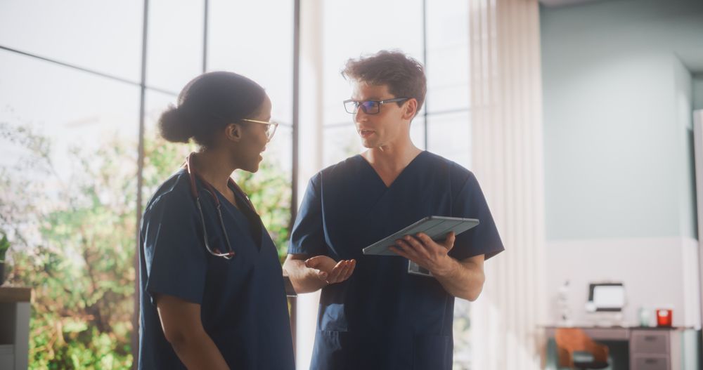 Two Young Doctors Standing in a Medical Facility in the Hospital, Talking and Sharing Information on Tablet Computer.