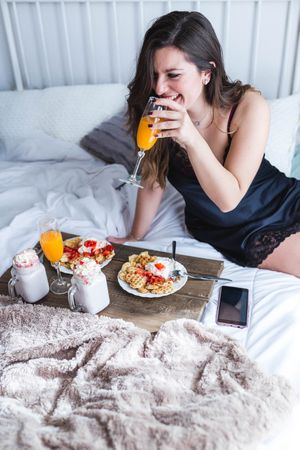 Young woman eating appetizing waffles and tasty drinks on tray on bed