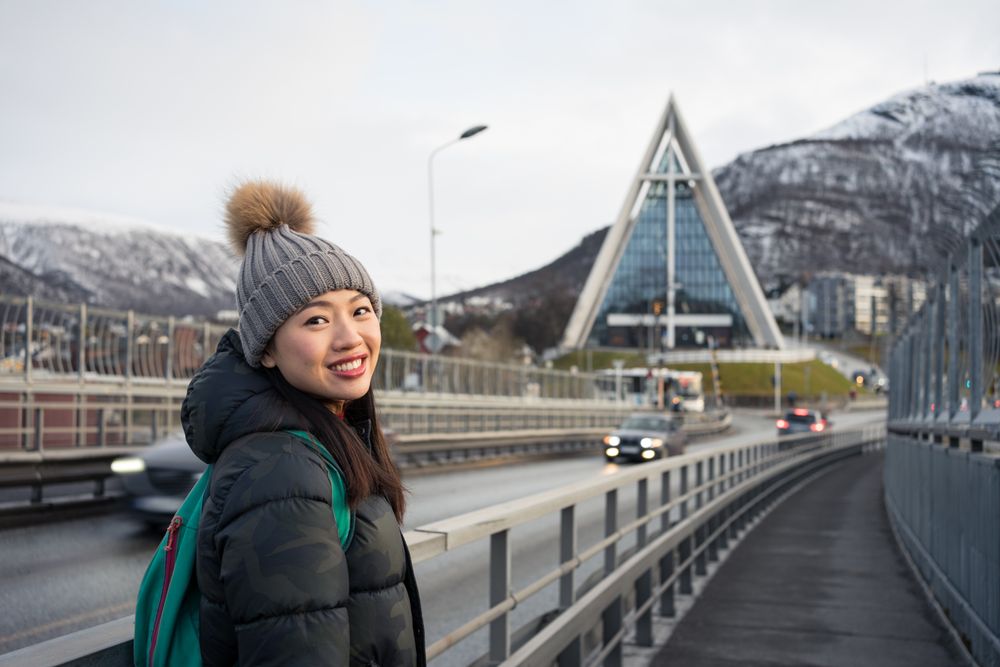 Joyful female tourist on road bridge against modern building with unusual architecture at foothill in cold weather