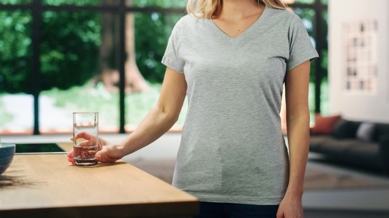 Young Adult Woman with Blond Hair Wearing Gray V-Neck T-Shirt, Holding a Glass of Water on the Table.