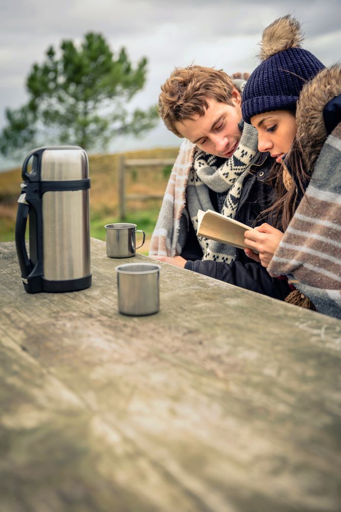 Couple sitting on park bench and making plans with flask of coffee on cold autumn day, vertical