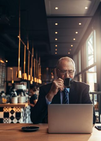 Mature man in business suit sitting at cafe table with laptop drinking coffee