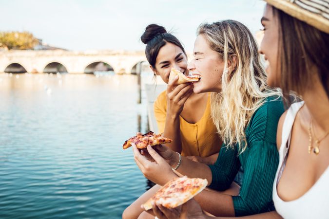 Side view of three happy female friends eating pizza on riverbank